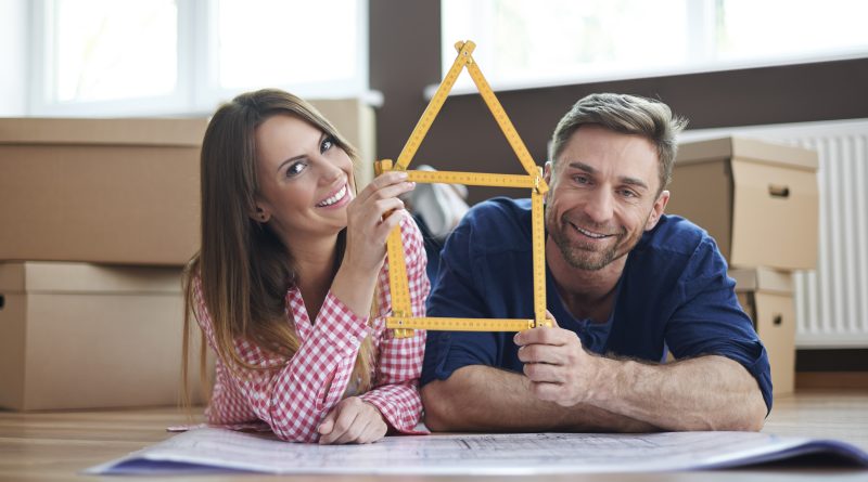 Uśmiechnięta para leży na podłodze w nowym mieszkaniu pośród kartonowych pudeł przeprowadzkowych. Trzymają wspólnie żółtą miarkę składaną, uformowaną w kształt domku. Przed nimi rozłożone są plany architektoniczne budynku.