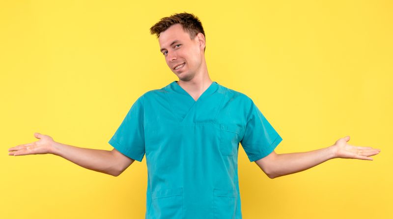 Młody mężczyzna w niebieskiej bluzie medycznej (scrubs) uśmiecha się i wykonuje gest rozłożonych rąk na jaskrawożółtym tle. Zdjęcie przedstawia profesjonalną męską odzież medyczną w przyjaznej, pozytywnej prezentacji.