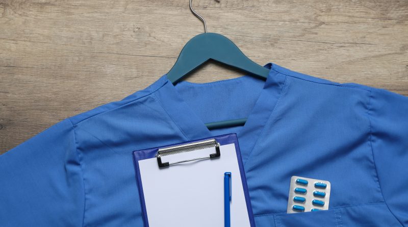 Niebieska bluza medyczna typu scrubs zawieszona na wieszaku, z przypiętym podkładem z klipsem oraz termoforem z tabletkami w kieszeni, na tle drewnianego blatu.