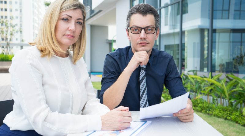 Kobieta i mężczyzna w strojach biznesowych siedzący przy stole z dokumentami, patrzący poważnie w stronę widza na tle nowoczesnego biurowca – symbol weryfikacji kontrahenta.