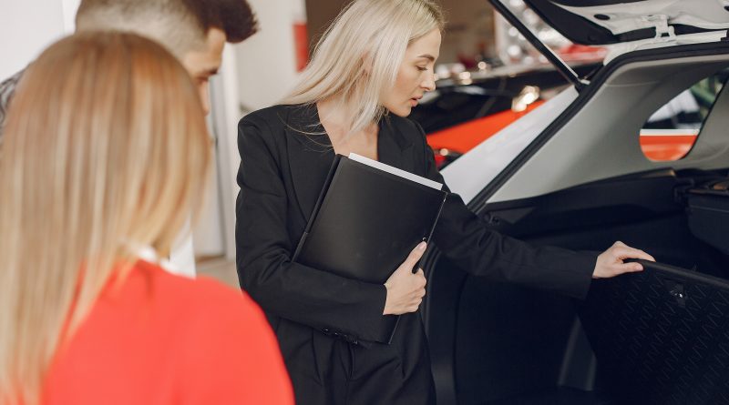 Blondwłosa kobieta w ciemnej marynarce z teczką prezentuje otwarty bagażnik samochodu parze klientów