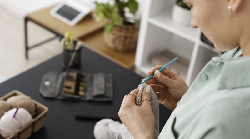 Kobieta ucząca się szydełkowania trzyma w dłoniach jasną włóczkę i niebieskie szydełko ergonomiczne