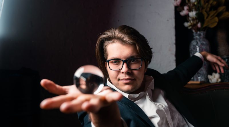 Iluzjonista w ciemnej marynarce i okularach prezentuje sztuczkę z przezroczystą szklaną kulą na wyciągniętej dłoni