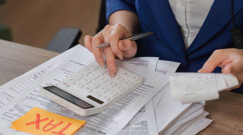Kobieta w niebieskiej marynarce licząca podatki na kalkulatorze i trzymająca paragon przy stosie dokumentów z żółtą karteczką TAX